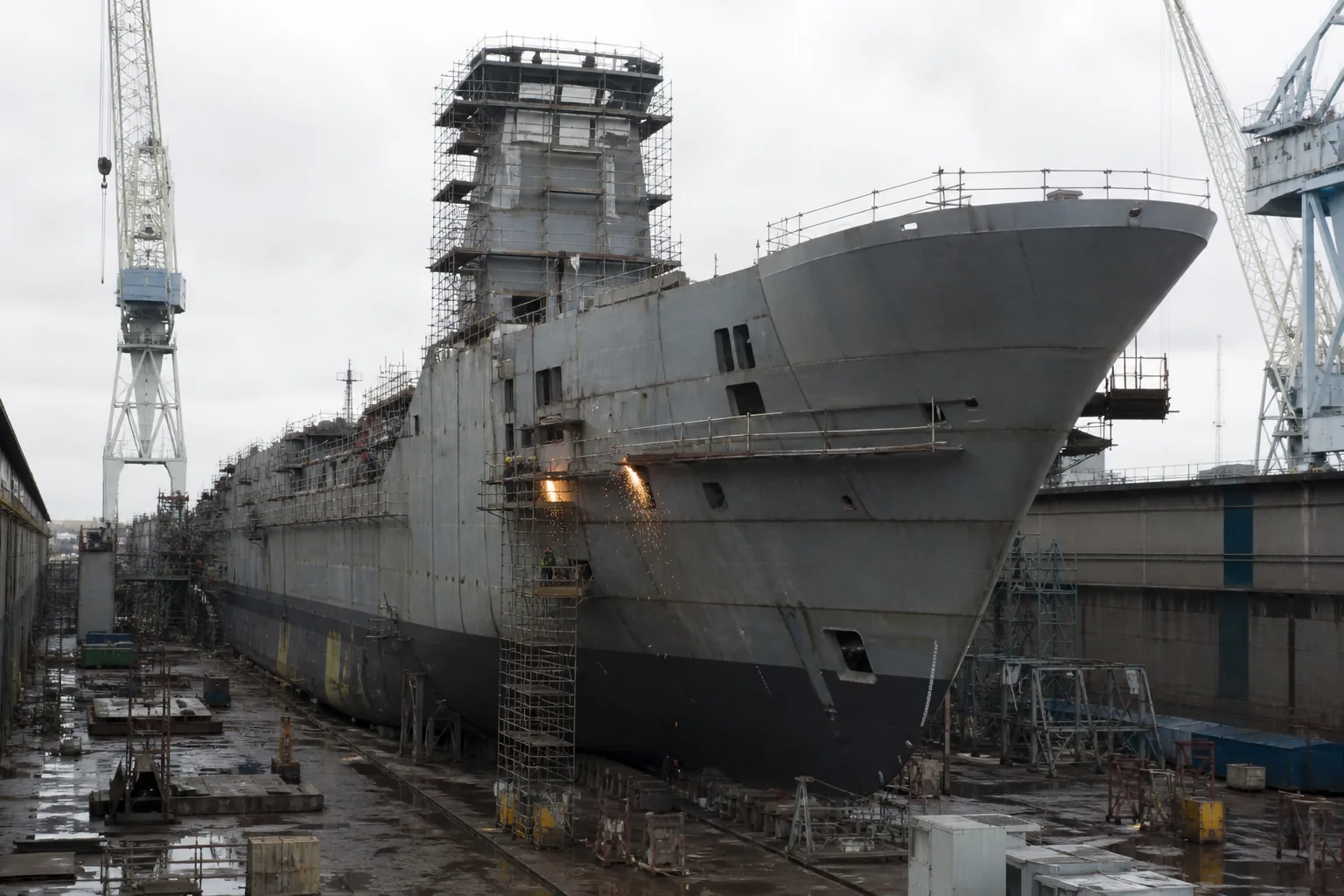 A very large grey ship hull under construction in a dry dock, surrounded by cranes and scaffolding (AI-generated image)