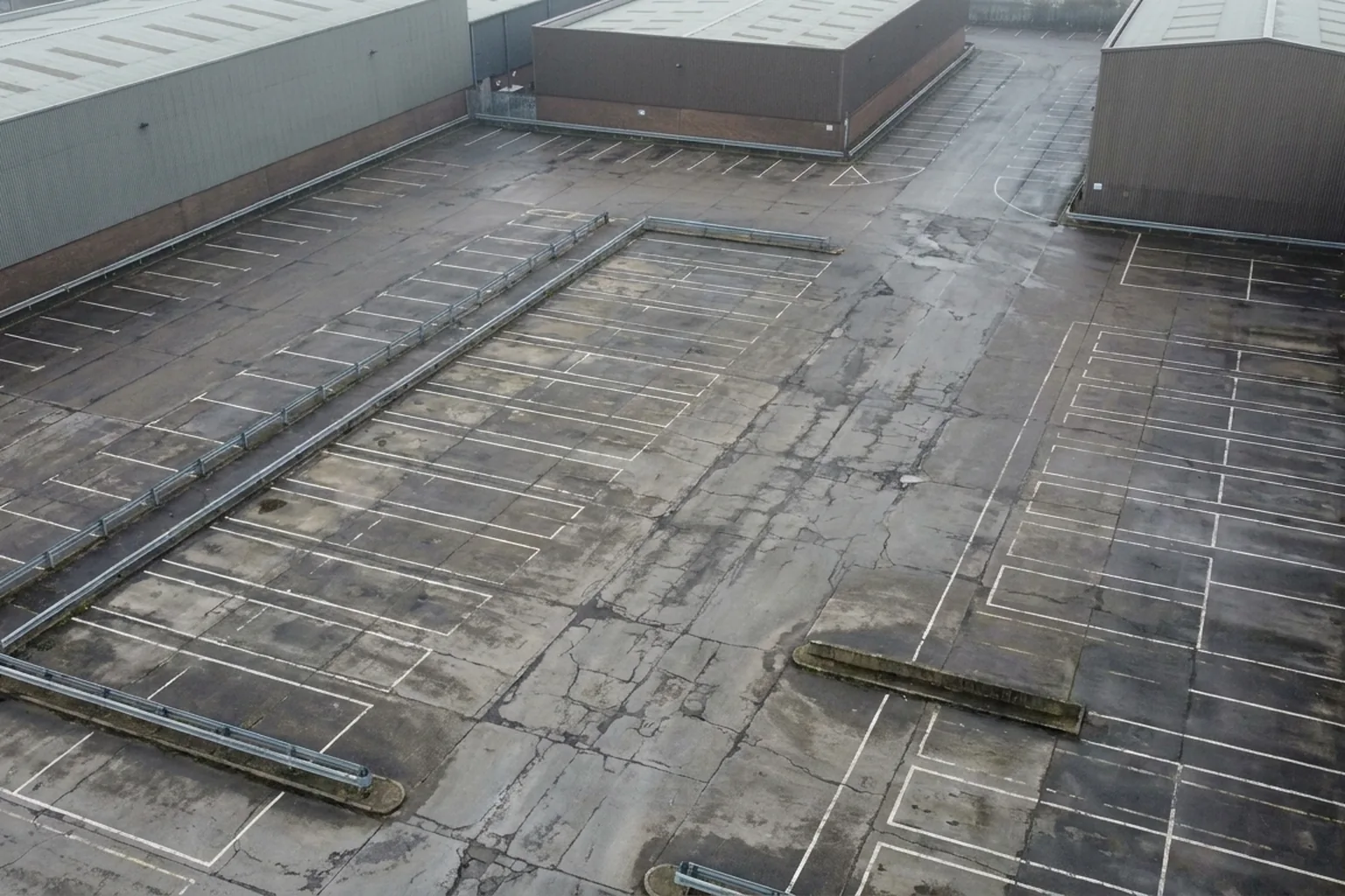 Empty, cracked asphalt parking lot marked with faded white lines and metal barriers, surrounded by drab industrial buildings (AI-generated image)