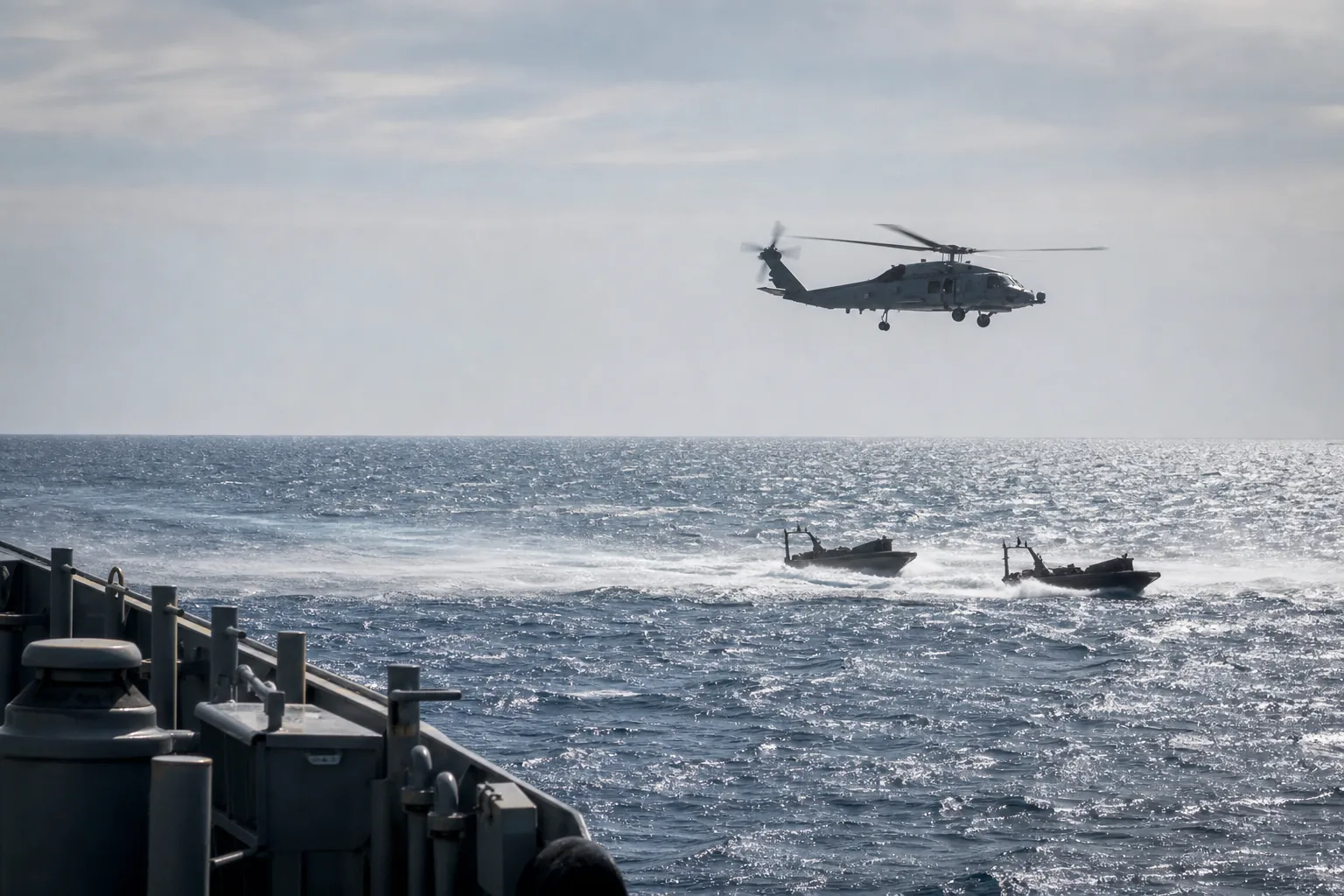 A naval helicopter hovering low over open ocean above two small open skiffs, seen from a ship at distance (AI-generated image)