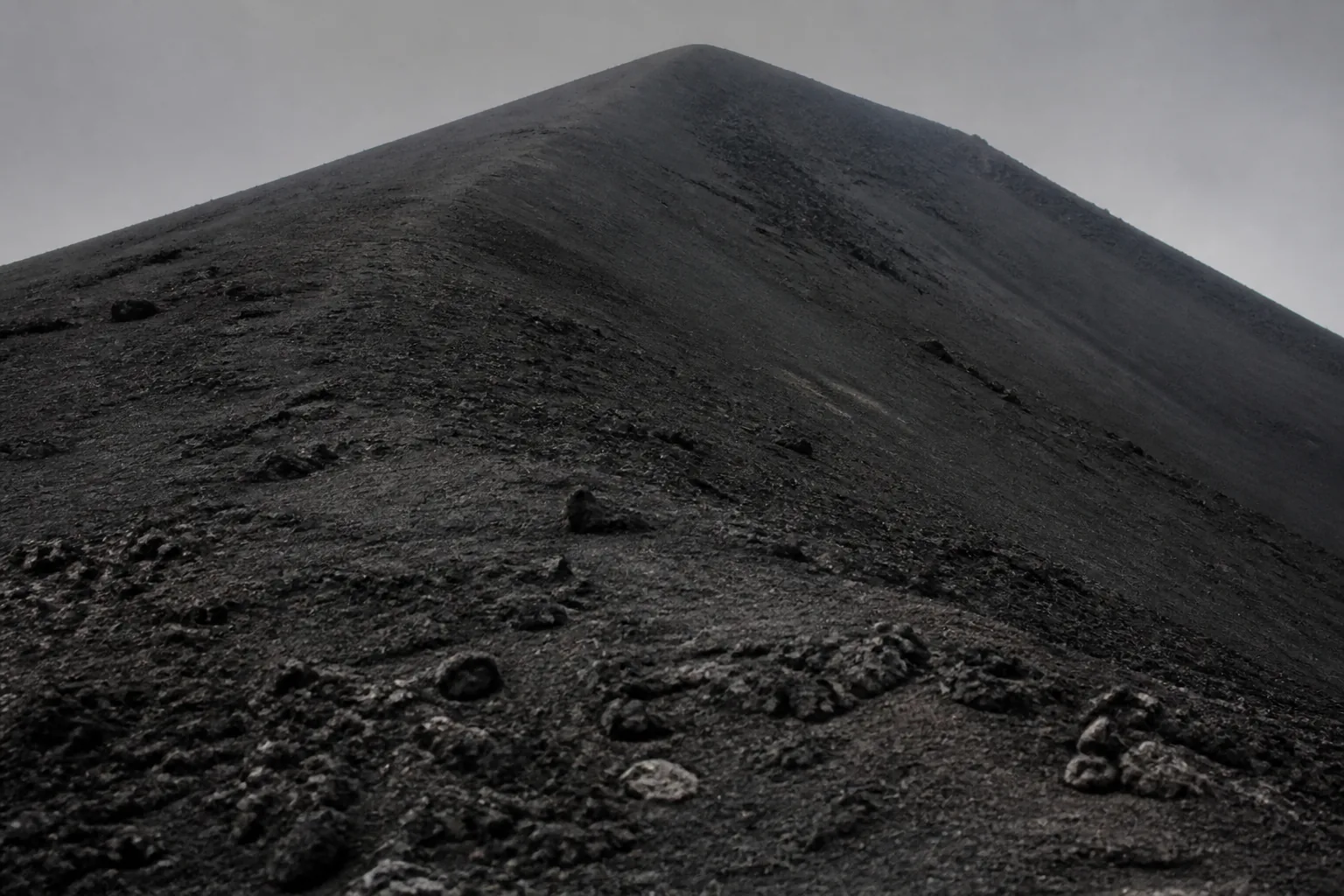 A black volcanic ash slope rising steeply to a bare summit, loose cinder underfoot (AI-generated image)