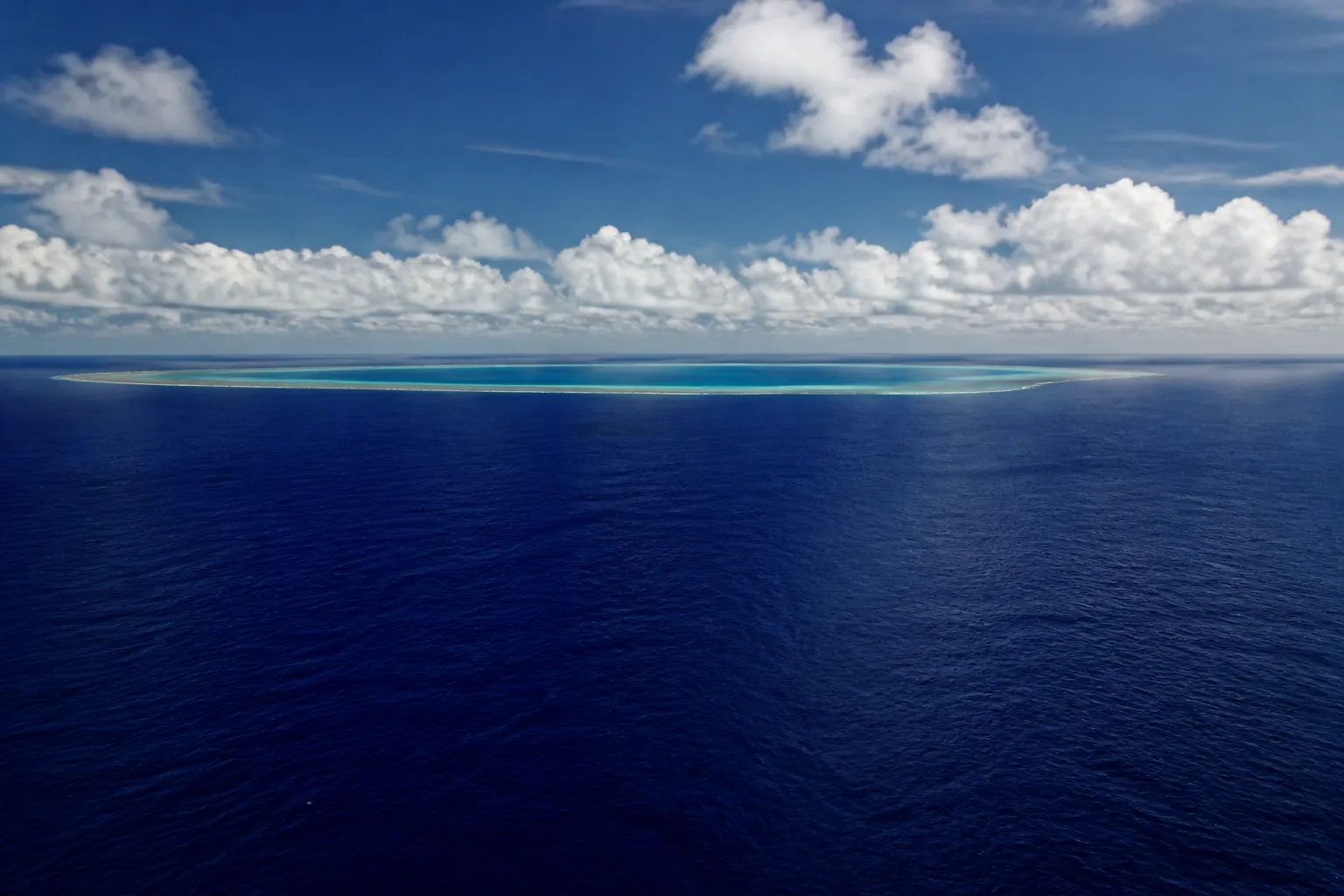 Open Pacific ocean from low altitude, deep blue water and a low coral atoll on the horizon (AI-generated image)