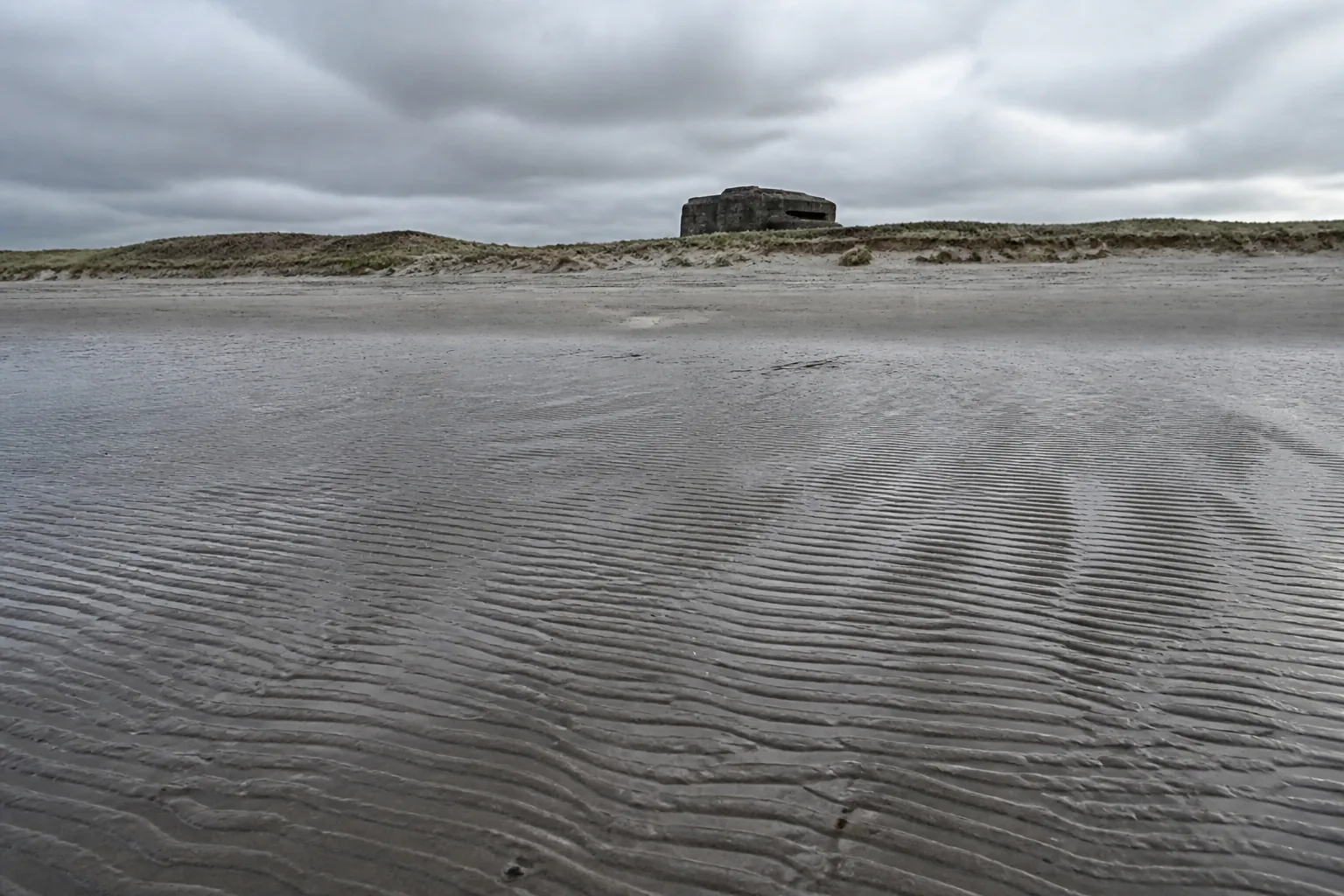 A wide flat beach at low tide with a concrete casemate on the distant dune line (AI-generated image)
