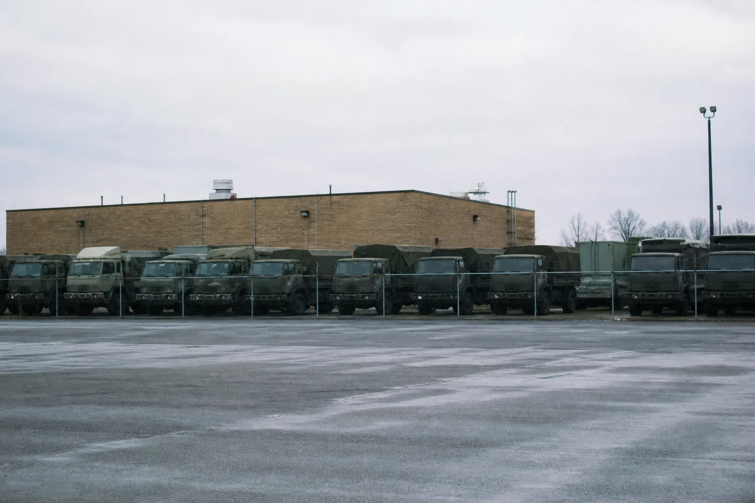 A line of military trucks parked in a fenced motor pool beside a plain brick facility (AI-generated image)