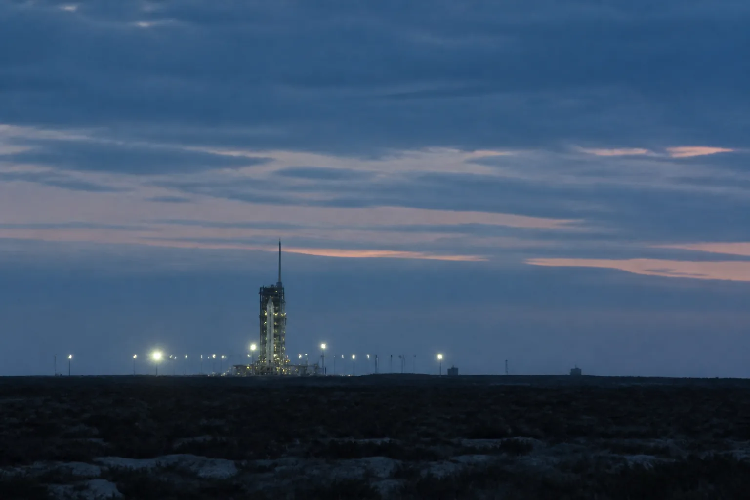A launch range at dusk: a distant service tower and floodlit pad seen across flat scrubland (AI-generated image)