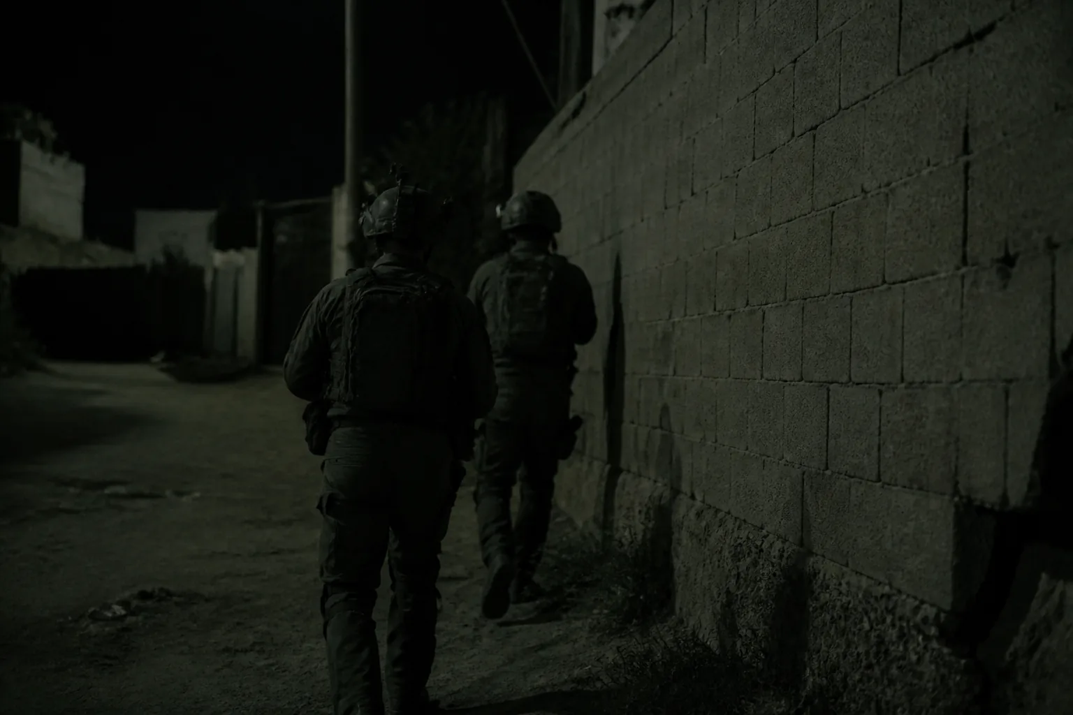 Two figures moving along a wall at night in low light, seen from behind at distance (AI-generated image)