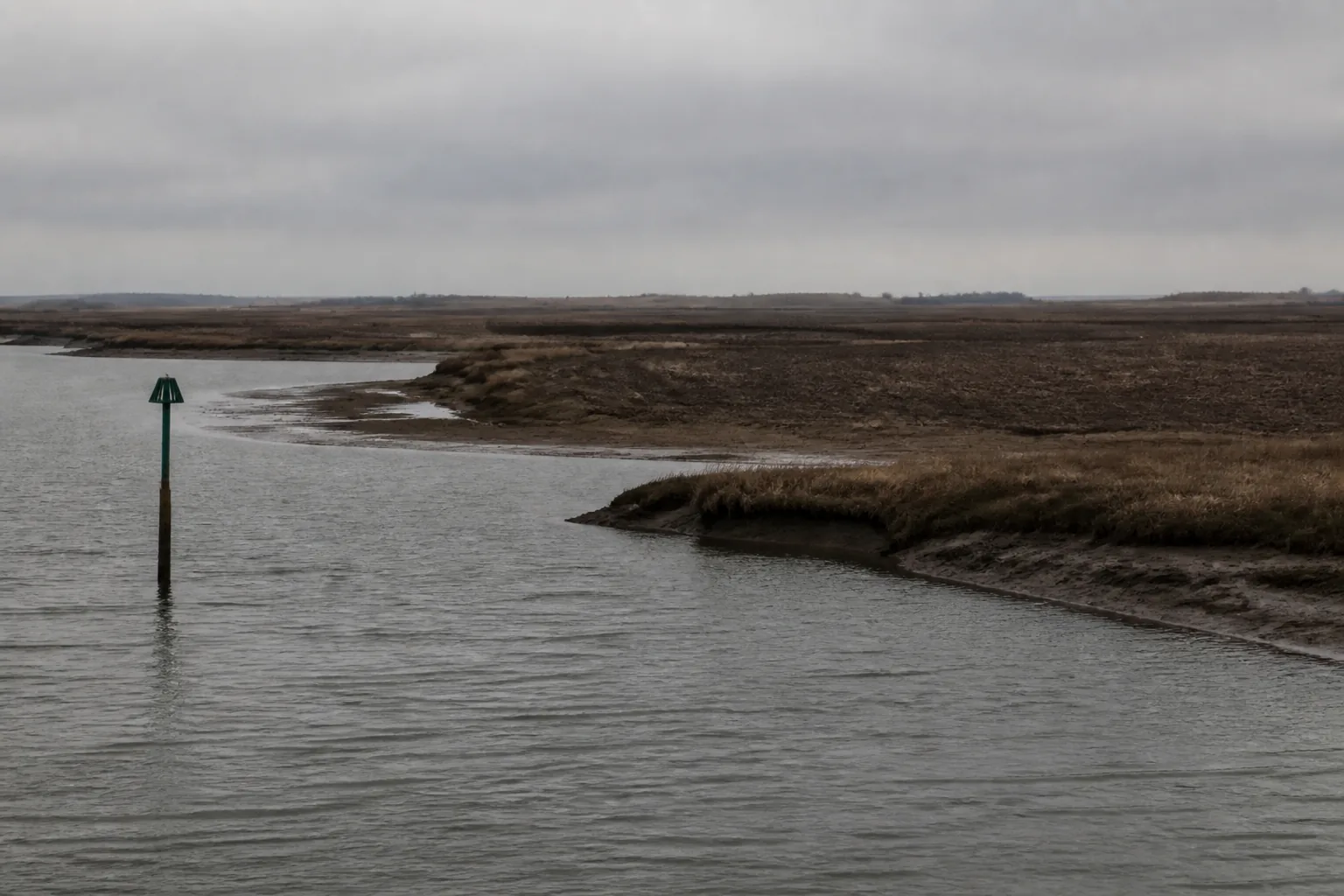 A river estuary where mudflats meet ploughed farmland, a channel marker standing in the water (AI-generated image)
