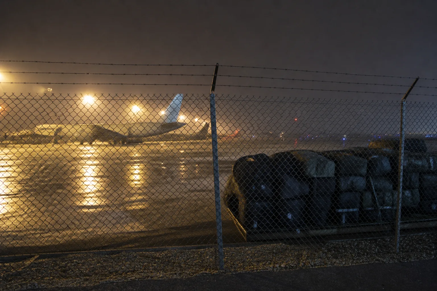An airfield departure area at night seen through a chain-link fence, kit bags stacked under floodlights (AI-generated image)