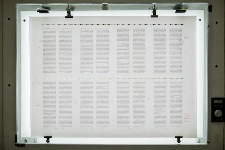 A printer’s proof sheet clipped to a light box, columns of type as grey blocks with red correction marks in the margins (AI-generated image)