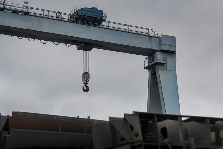 A shipyard gantry crane against an overcast sky with its hook block lowered over stacked hull sections (AI-generated image)