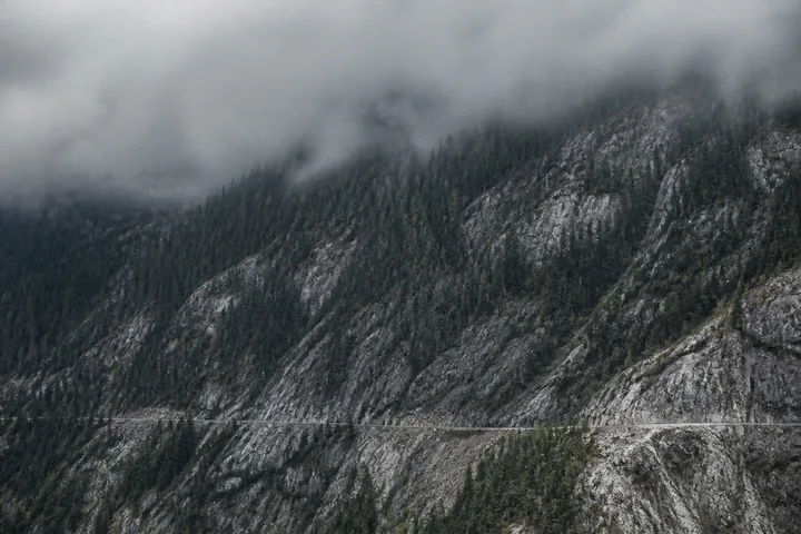 A forested granite mountainside with a service road cut into it under heavy grey cloud (AI-generated image)