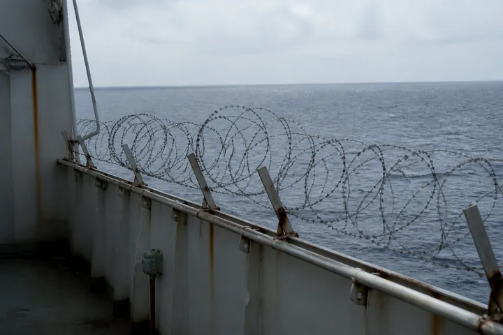 A merchant ship’s rail wrapped in razor wire with the open ocean beyond in hazy tropical light (AI-generated image)