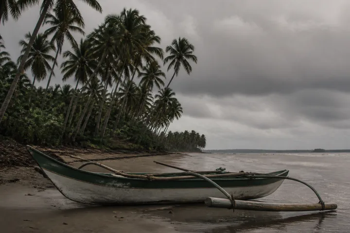A small fishing outrigger canoe pulled up on wet sand under heavy grey monsoon cloud, palms leaning behind it (AI-generated image)