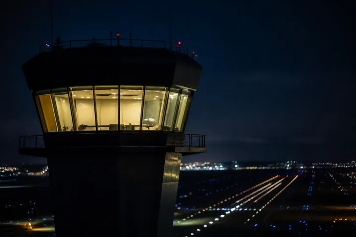 An air traffic control tower at night, its glass lit from within, runway lights receding into the darkness (AI-generated image)