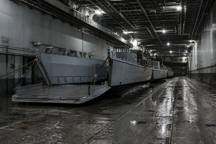 A ship’s well deck interior with landing craft chained down, water sheened on the deck under harsh work lighting (AI-generated image)