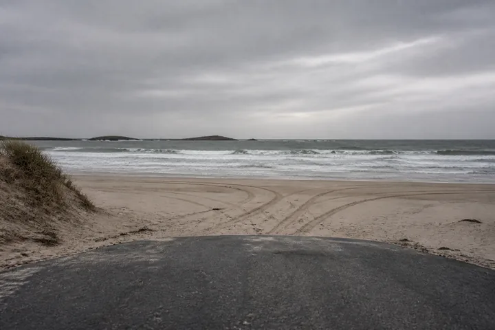 A beach where a metalled road ends at the sand, tyre tracks stopping at the tideline under overcast light (AI-generated image)