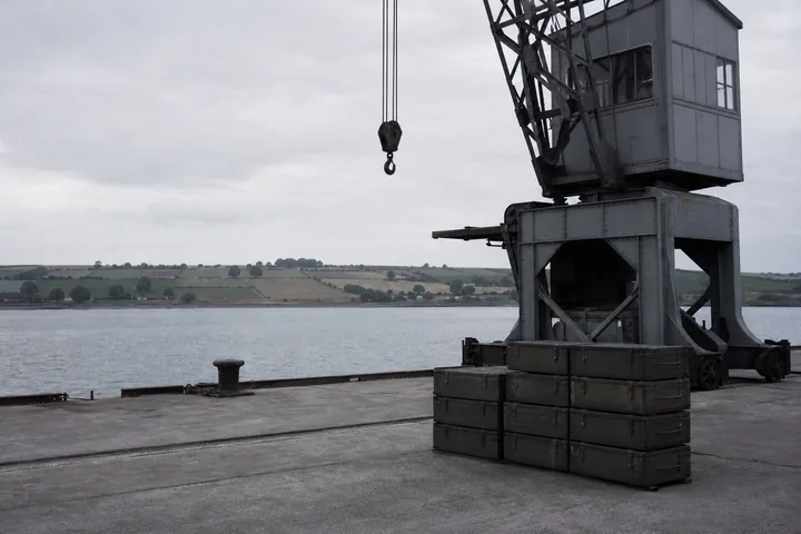 A dockside crane and stacked crates on a quay with farmland visible across the water (AI-generated image)