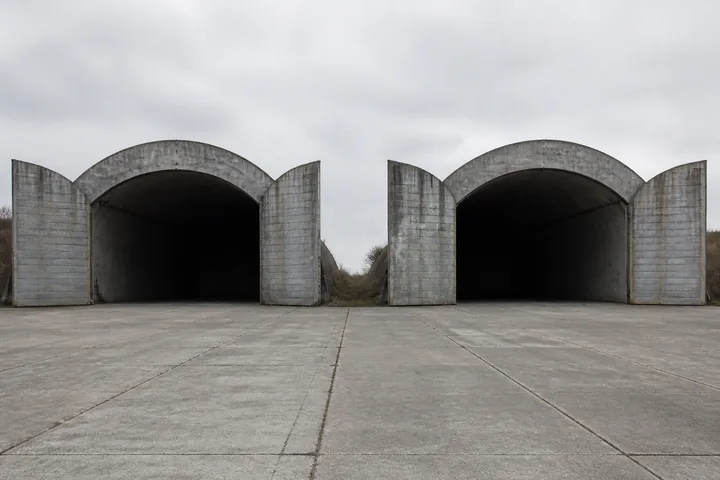 Two empty hardened aircraft shelters side by side on a concrete apron, doors open and interiors dark (AI-generated image)