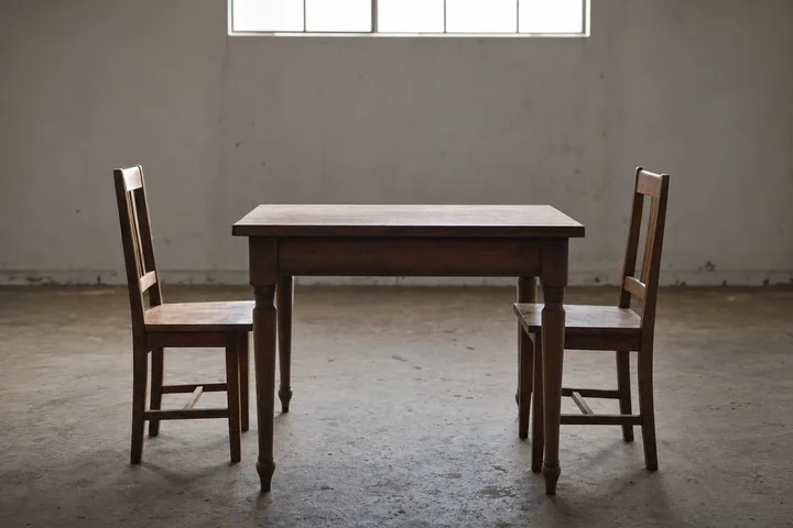 Two plain wooden chairs facing each other across an empty table in a bare room, hard light from a high window (AI-generated image)