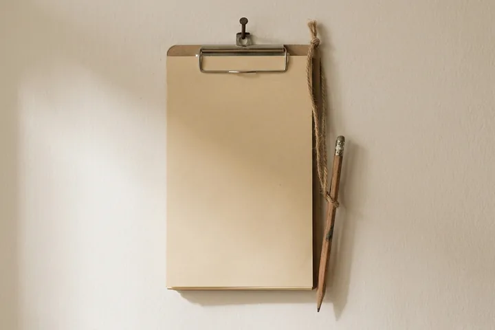 A brown clipboard with blank beige paper and a tied wooden pencil hanging on a light beige wall (AI-generated image)