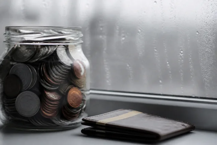 A jar of coins and a folded bank card on a windowsill with rain on the glass behind (AI-generated image)