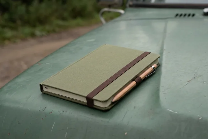 Closed olive-green notebook with a brown elastic securing a wooden pencil, on a scratched dark green vehicle hood (AI-generated image)