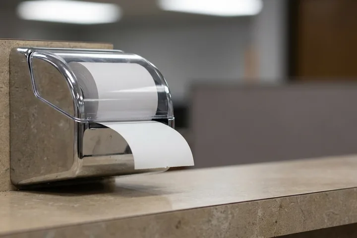Chrome and clear plastic dispenser with white paper unrolled, mounted on a speckled wall above a matching countertop (AI-generated image)