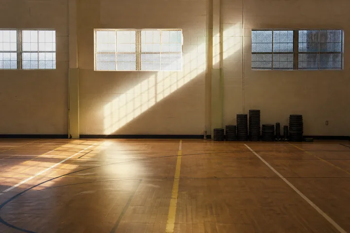 A gymnasium floor with painted line markings and a stack of weight plates against the wall under high windows (AI-generated image)