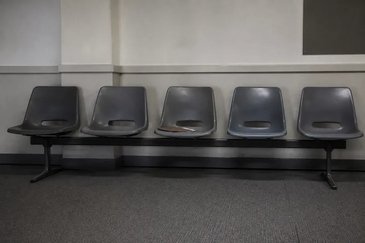 A row of plastic waiting-room chairs bolted to a rail in a government office, a clipboard left on one seat (AI-generated image)