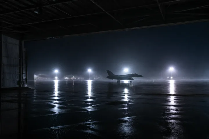 The view out of an open hangar onto a rain-soaked apron at night, floodlights reflected in the puddles (AI-generated image)
