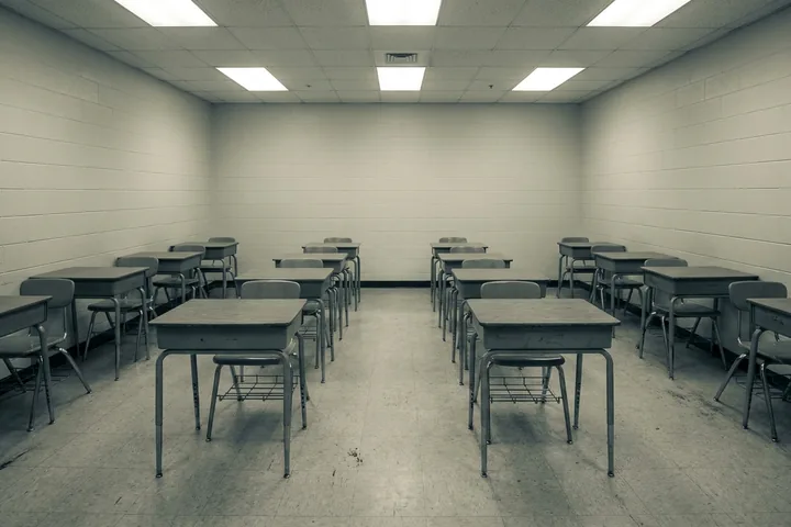 Empty rows of student desks and chairs arranged in a grey, block-walled classroom (AI-generated image)