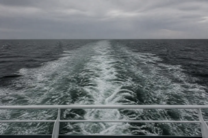 A wake spreading behind a ship, seen from the stern rail, grey swell to the horizon (AI-generated image)