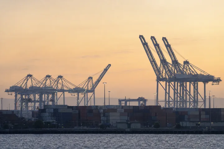 A container port at dawn seen from a distance, stacked boxes and gantry cranes in silhouette (AI-generated image)
