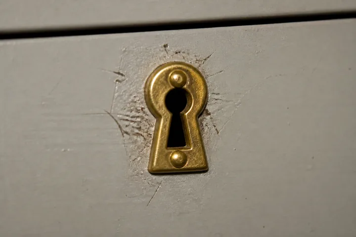 A small brass keyhole escutcheon on a plain painted metal drawer front, scratched around the opening (AI-generated image)