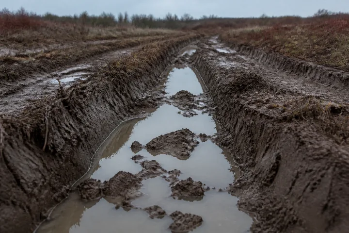 Deep track ruts cut through wet clay on a training area, water standing in them under grey light (AI-generated image)