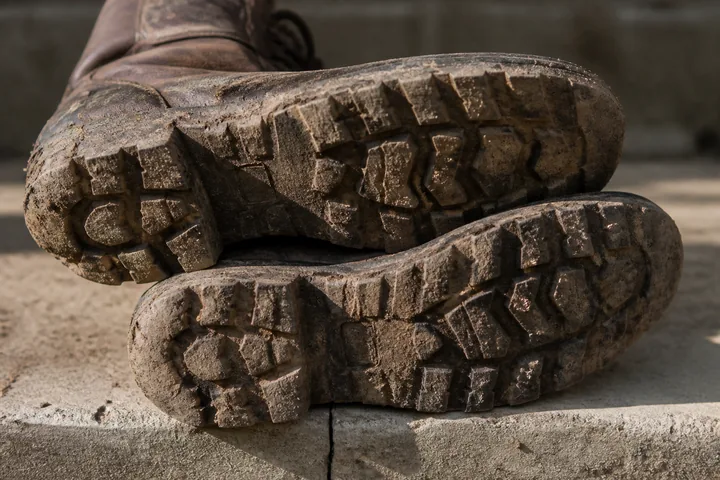 Worn boot soles caked in dried mud resting on a concrete step, tread detail picked out by hard side light (AI-generated image)