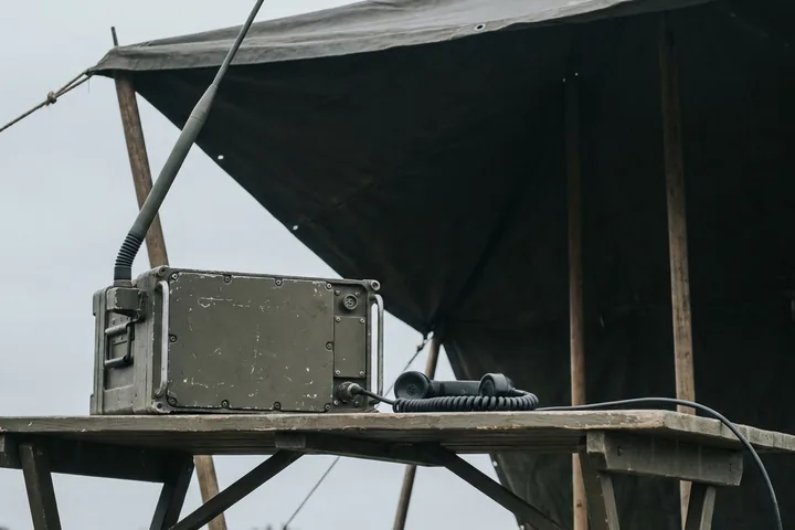 An olive green military radio with an antenna and telephone handset on a wooden field table under a canvas awning (AI-generated image)