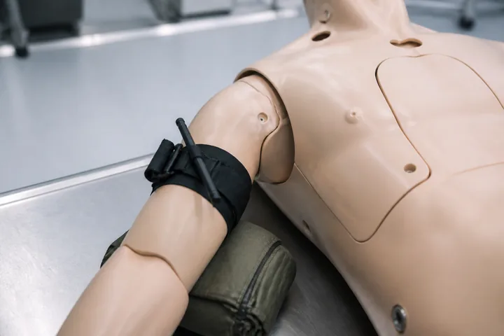 A training mannequin torso on a table with a tourniquet applied to a simulated limb under clinical lighting (AI-generated image)