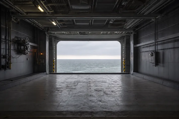 An aircraft elevator platform at hangar-deck level, its opening onto daylight and the sea (AI-generated image)