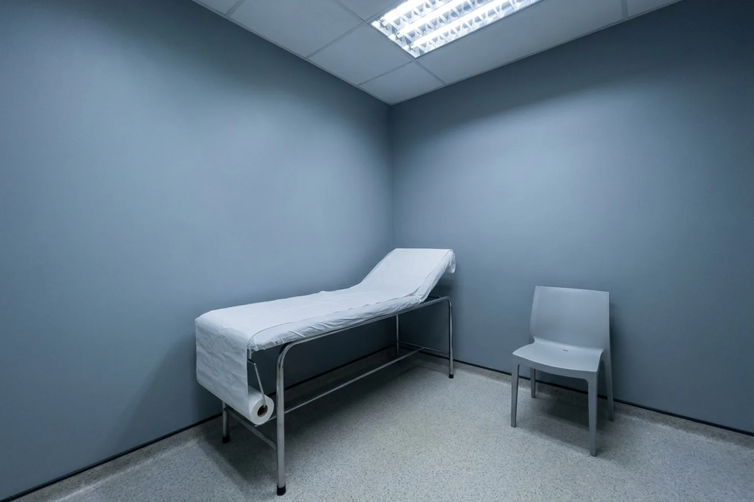 An examination table with white paper roll and grey chair in a blue-grey room with speckled floor and bright fluorescent light (AI-generated image)