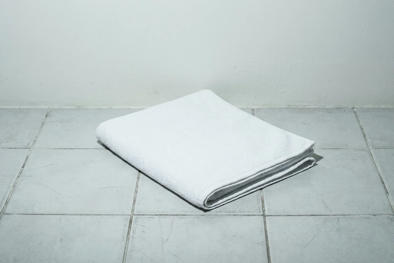 Neatly folded white towel casting a shadow on worn grey tiled floor against a plain light wall (AI-generated image)