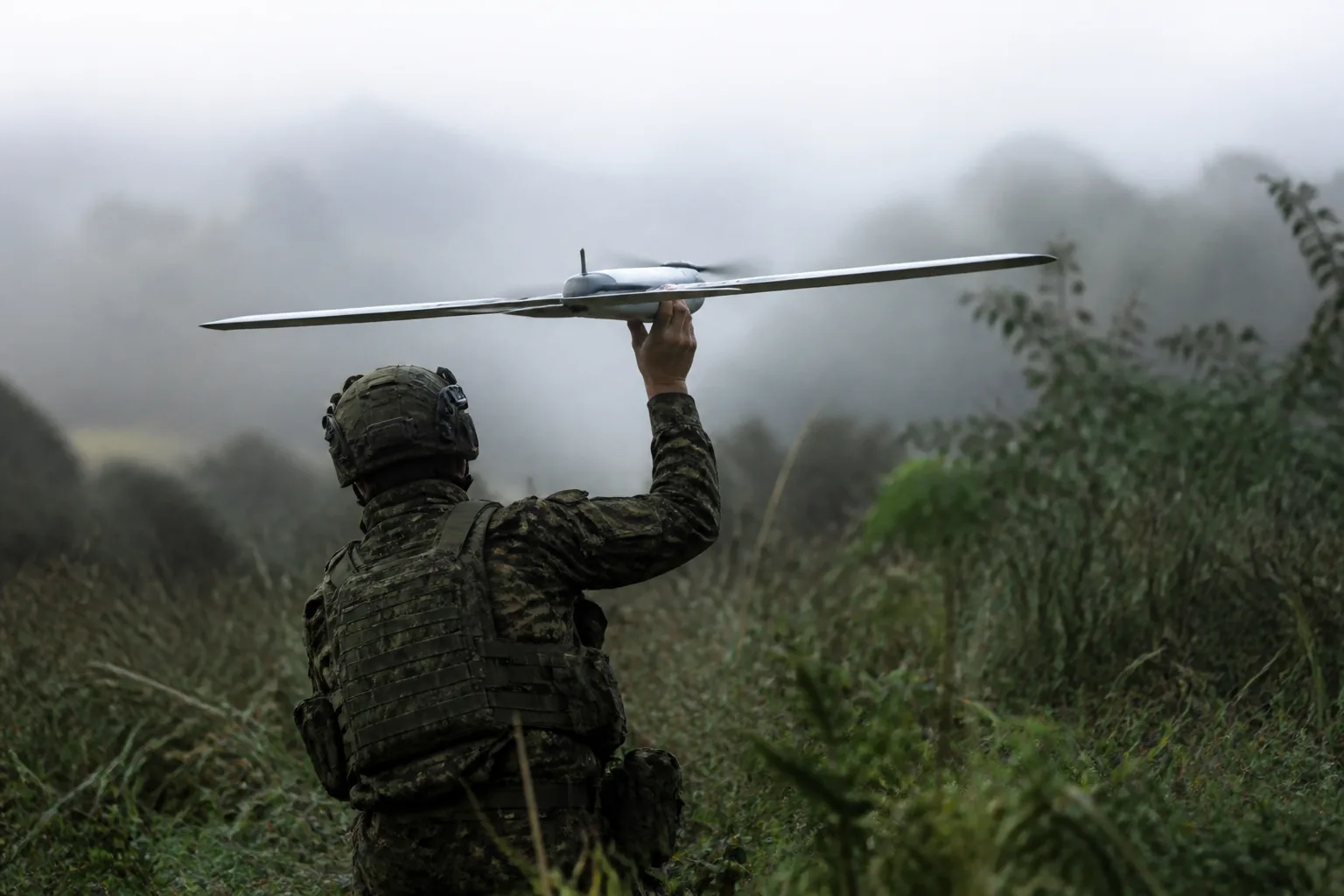 A soldier crouched in tall grass, holding a small reconnaissance drone above his head (AI-generated image)
