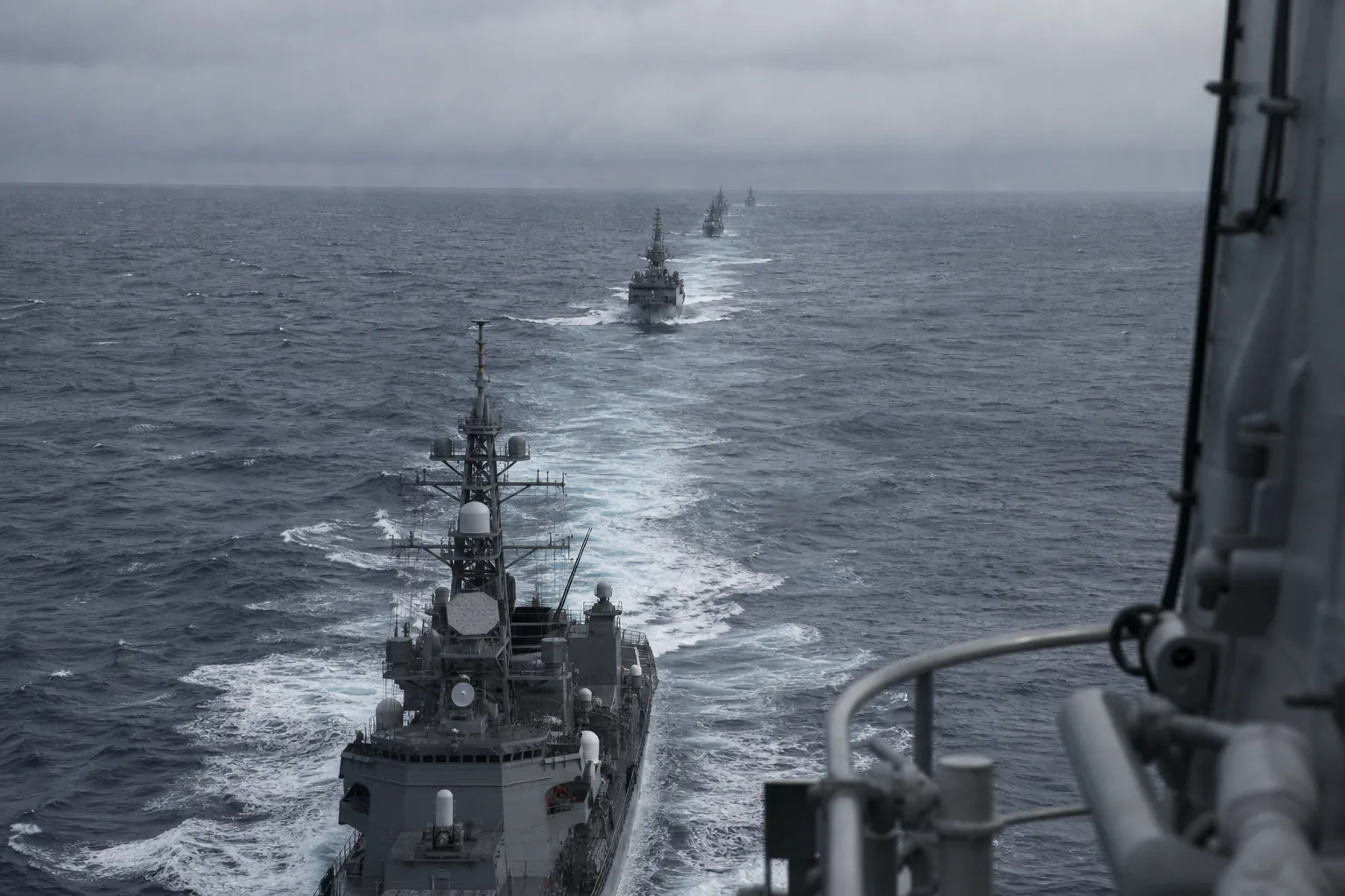 A line of grey warships under way in formation on open ocean, seen from a mast looking aft along the column (AI-generated image)