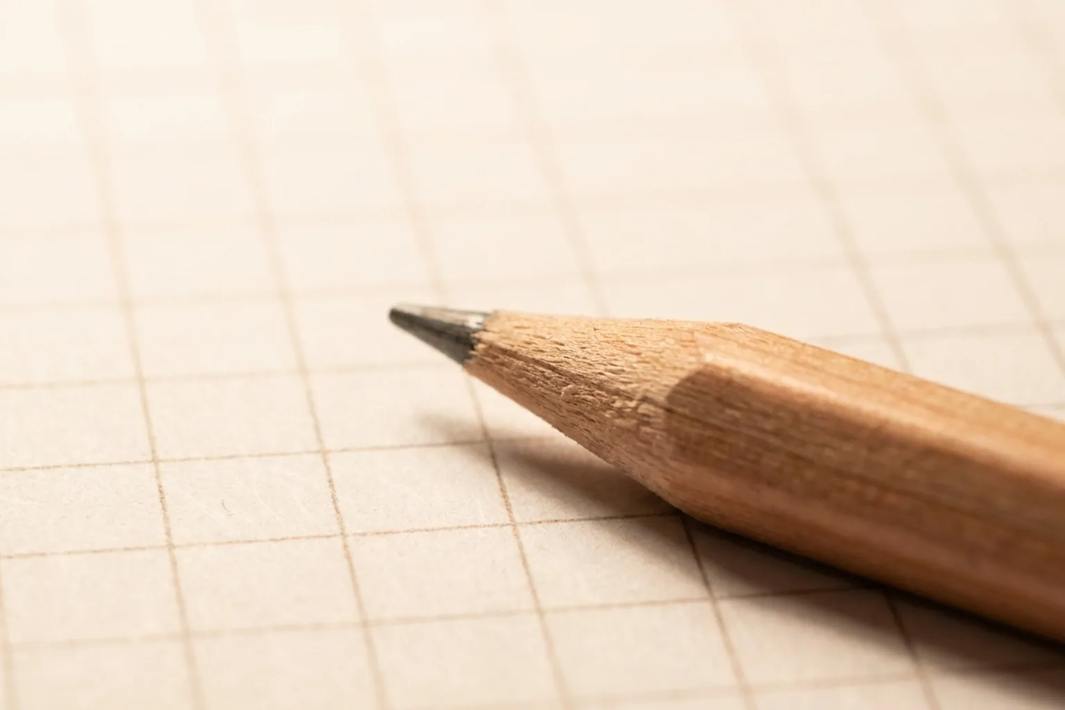 Sharpened light brown wooden pencil point in close-up, angled toward the upper left on beige grid paper (AI-generated image)