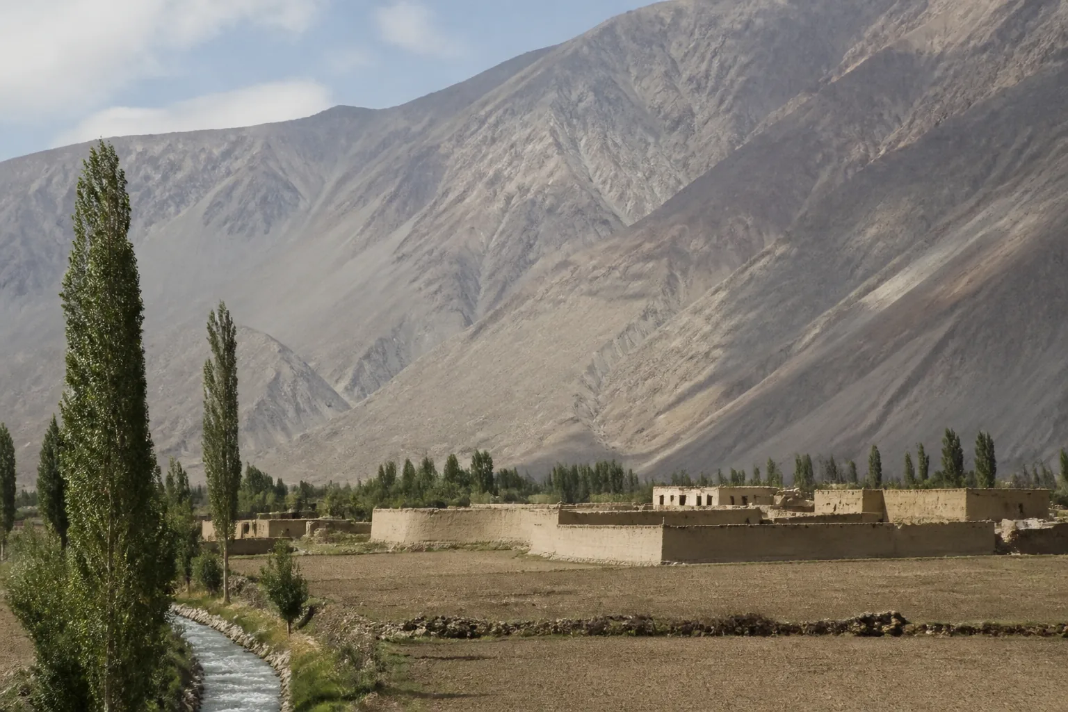 A mud-walled compound on a valley floor beneath enormous barren mountains (AI-generated image)
