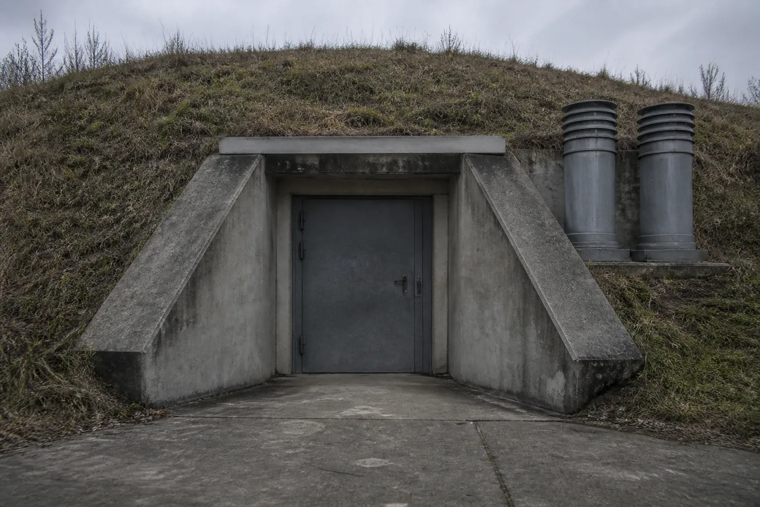 A hardened bunker entrance set into a grass bank, blast door closed, ventilation stacks beside it (AI-generated image)