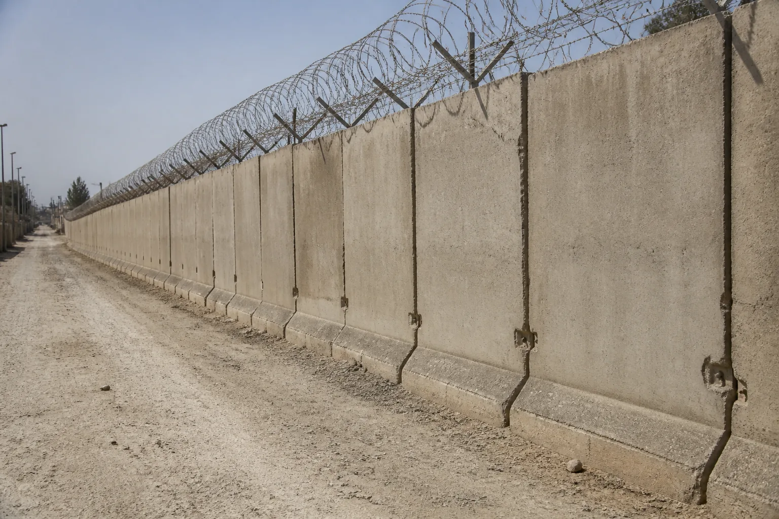 A corridor of interlocked concrete blast walls on a dusty street, razor wire above (AI-generated image)